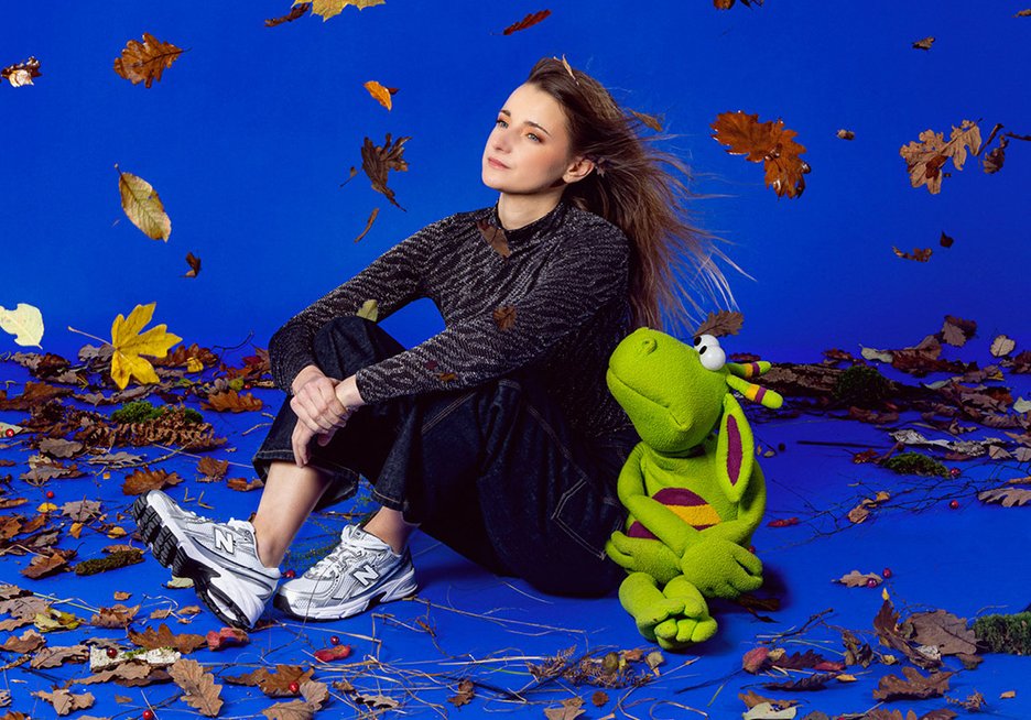 Capucine Duchamp assise au milieu de feuilles d'automne sur fond bleu avec Eliott, sa marionnette de dragon vert