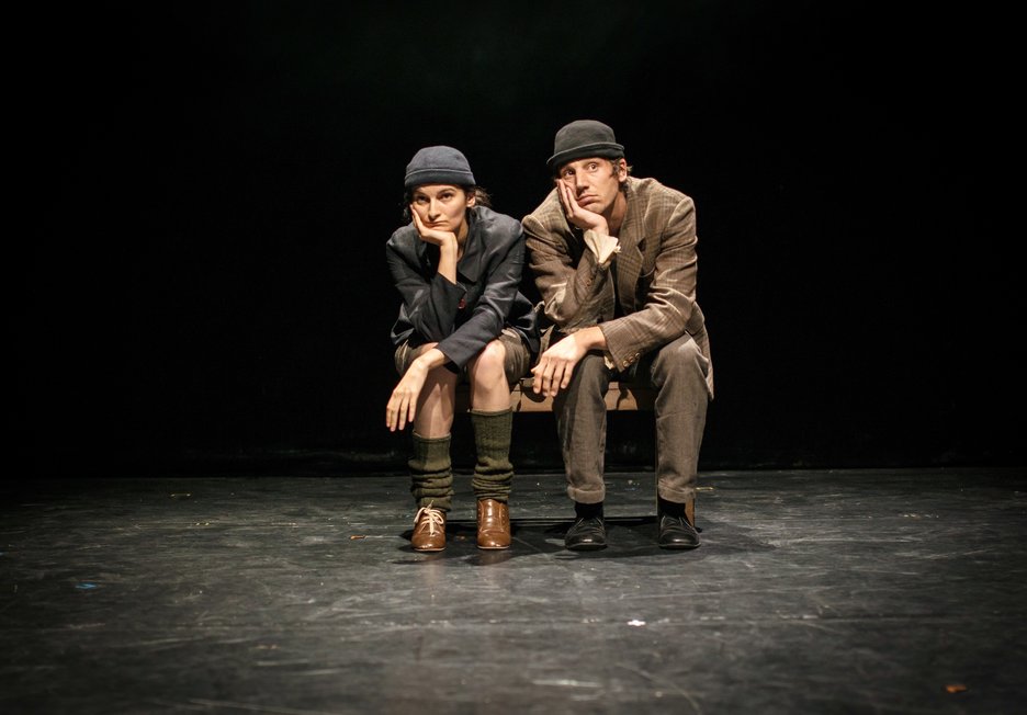 Deux comédiens assis sur un banc, en costume et chapeau, le menton dans la main, sur une scène sombre