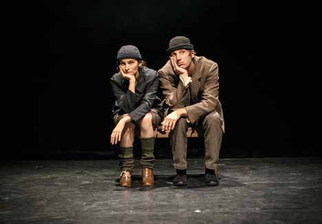 Deux comédiens assis sur un banc, en costume et chapeau, sur une scène sombre